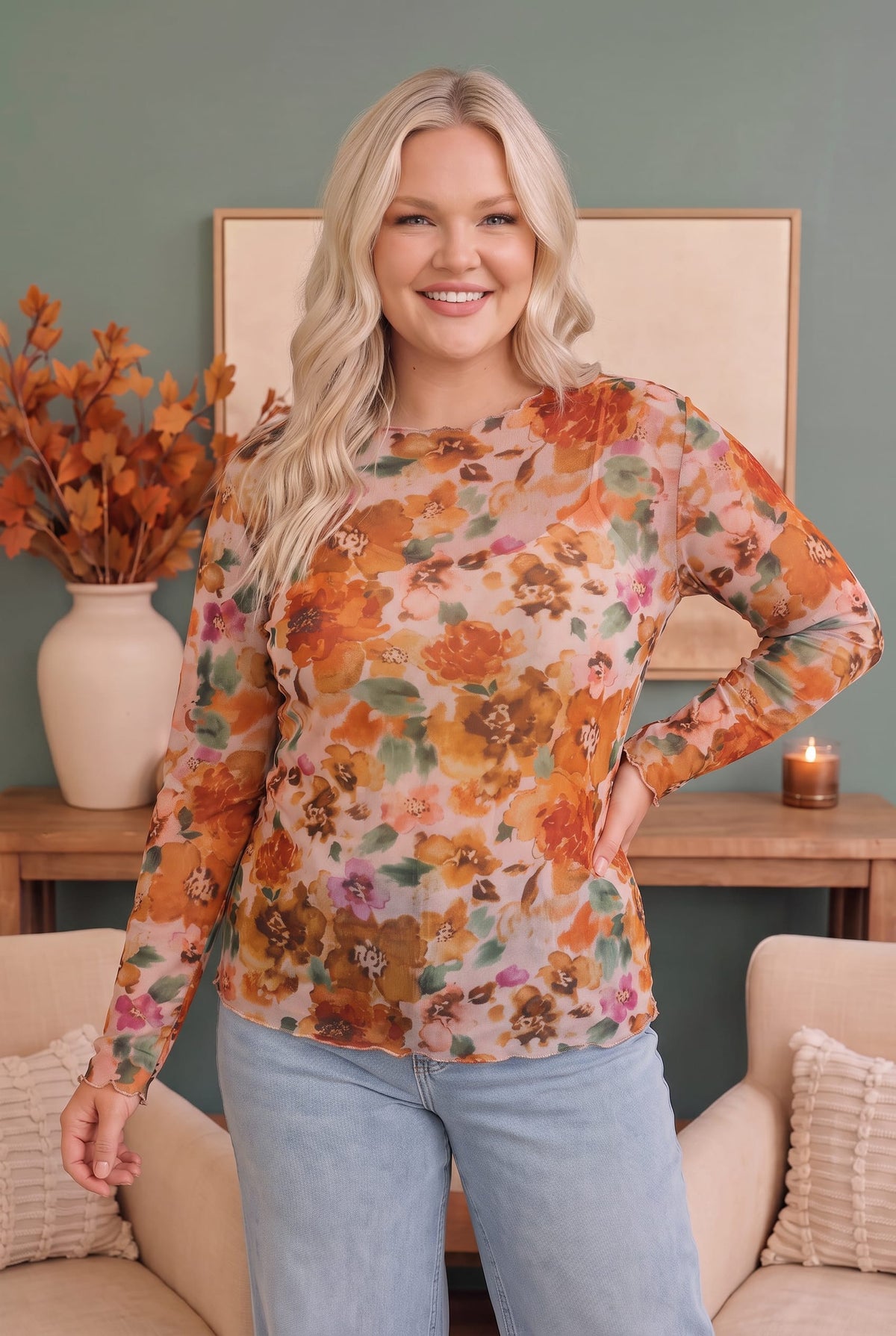 A woman modeling a long-sleeve floral top with a warm orange, brown, and green floral pattern on a beige background, paired with light blue jeans in a cozy living room setting.