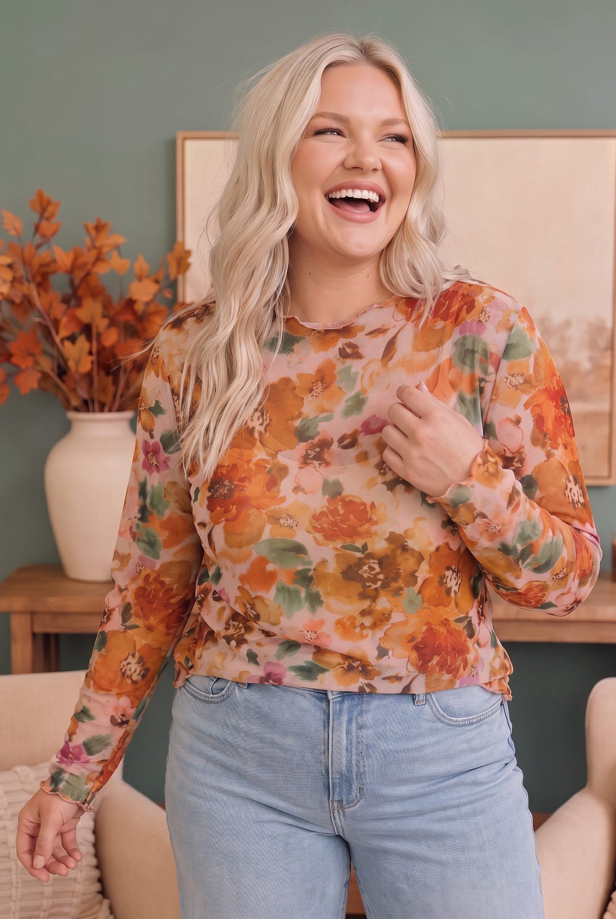 A woman with blonde hair wearing a long-sleeve floral print top in orange, yellow, and green tones, paired with light blue jeans, standing in a cozy room setting.