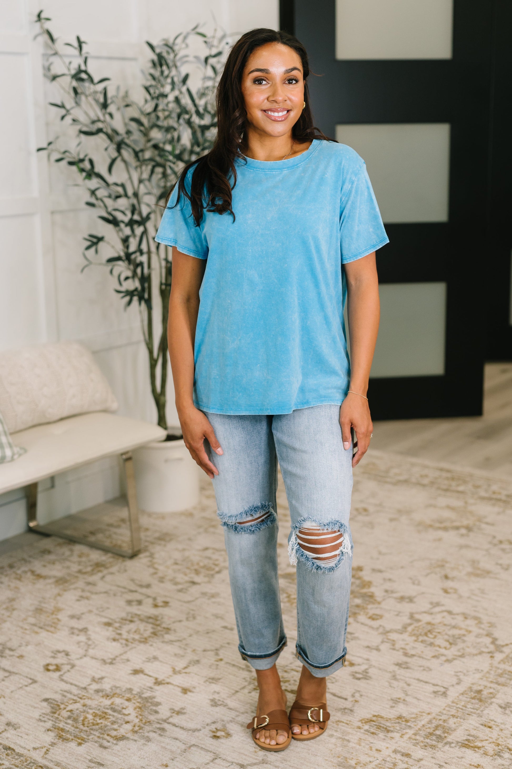Woman wearing a blue t-shirt and ripped jeans standing in a room with a plant and furniture.