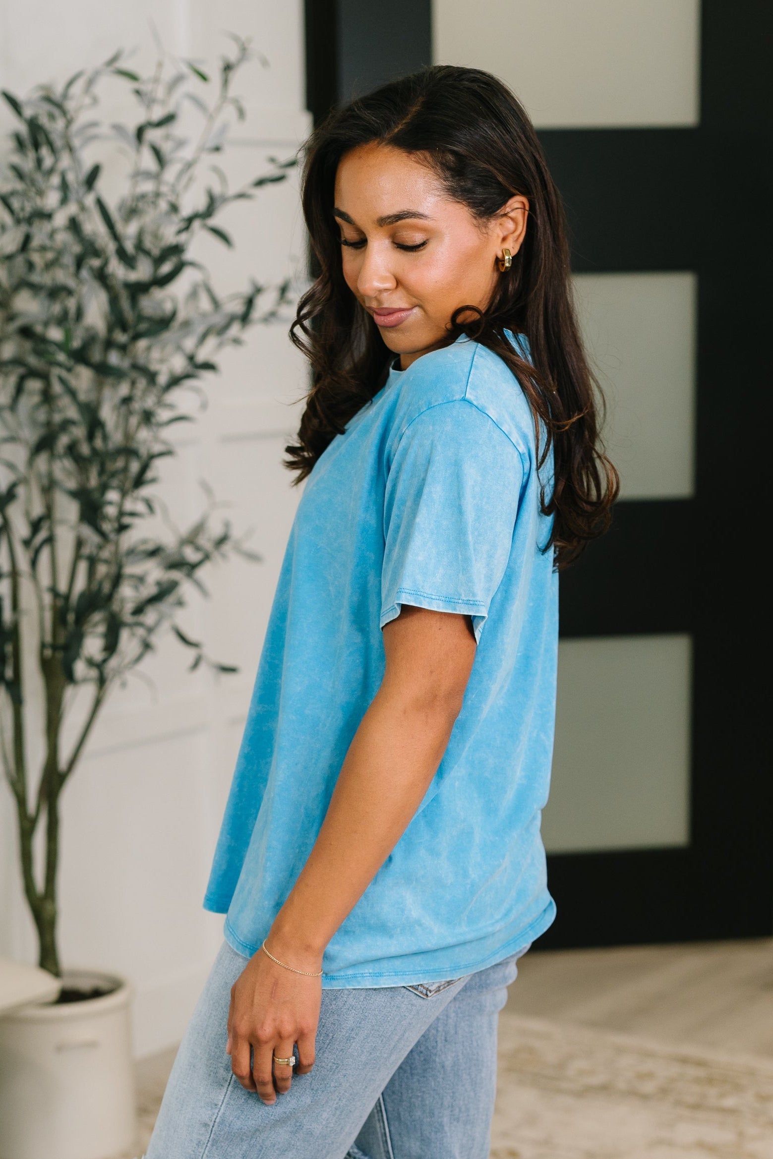 Woman wearing a light blue t-shirt and jeans standing indoors with a plant in the background