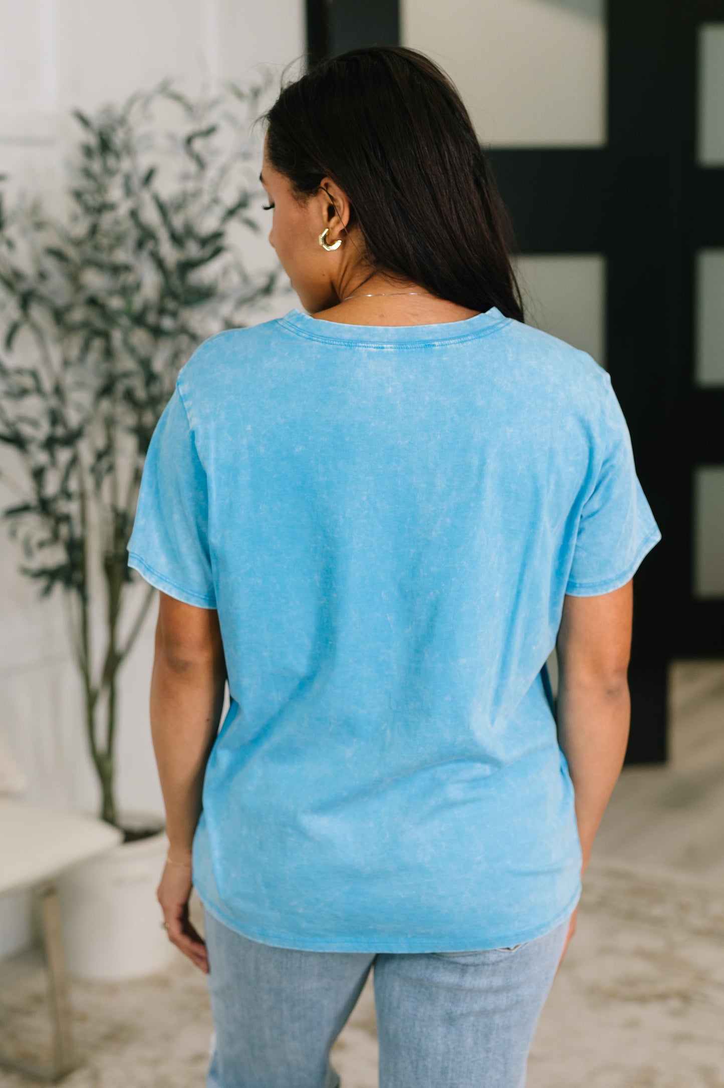 Woman wearing a light blue t-shirt and jeans standing indoors with a plant in the background