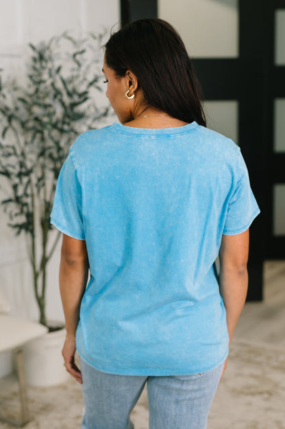 Woman wearing a light blue t-shirt and jeans standing indoors with a plant in the background