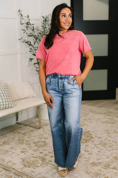 Woman wearing a pink t-shirt and blue jeans standing in a room with a plant and bench.