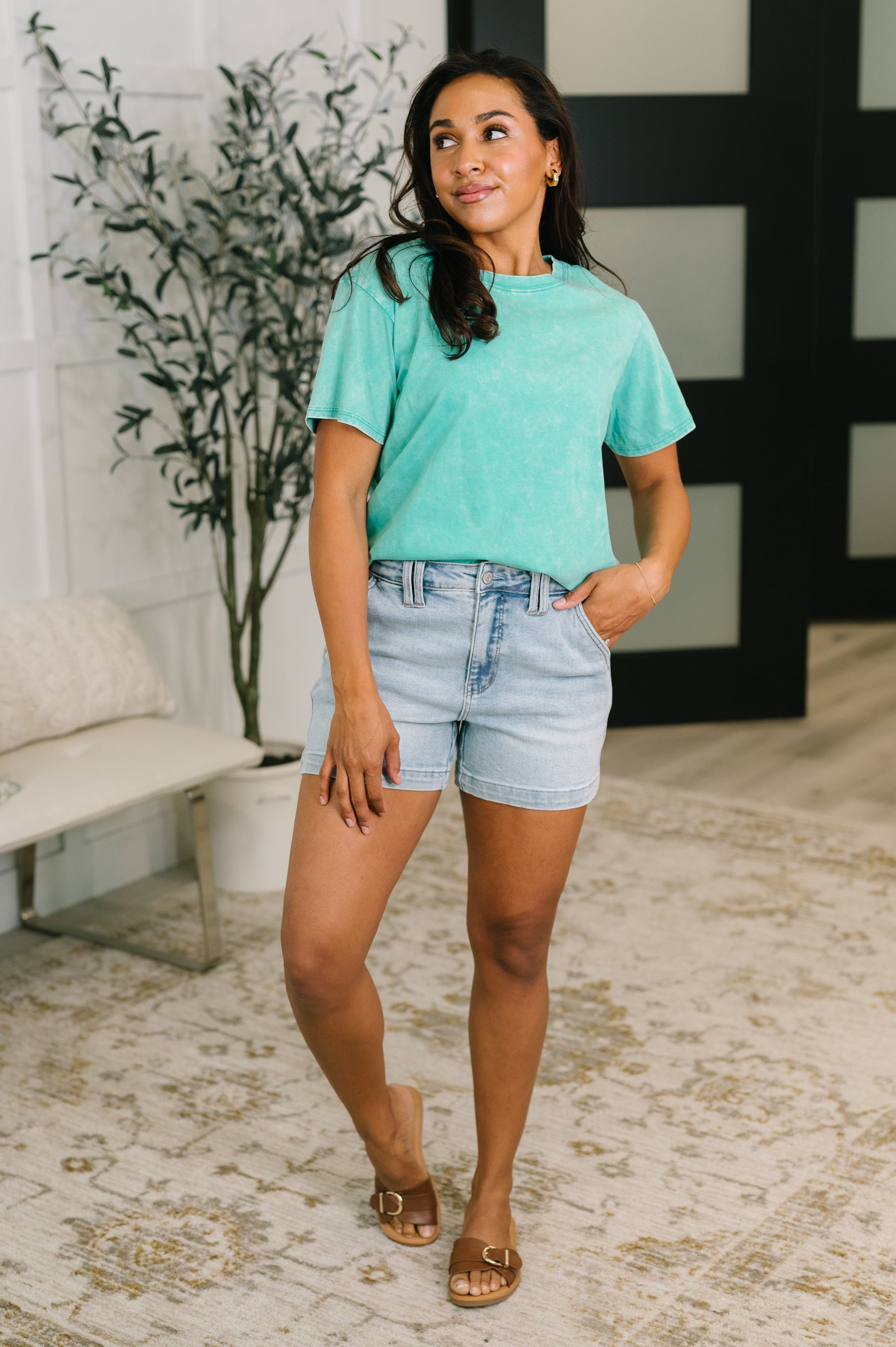 Woman in a teal shirt and denim shorts standing in a room with a plant and bench.