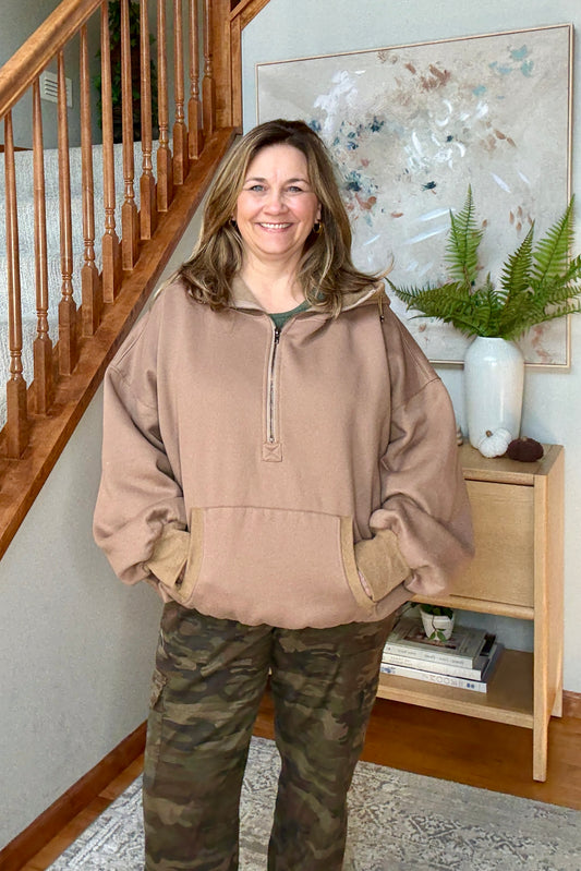 Woman wearing a brown hoodie 1/2 zip sweatshit and camouflage pants standing in a home setting with a staircase and decor.