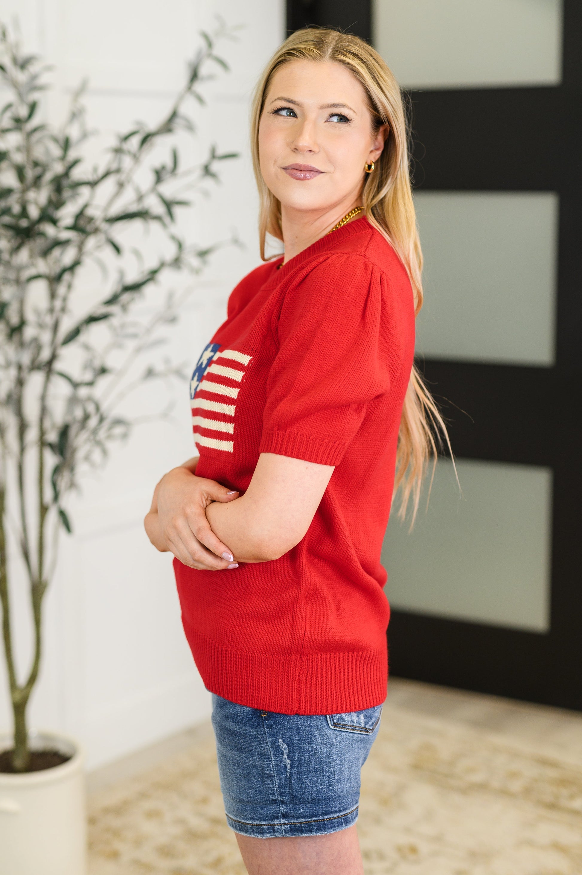Woman wearing a red sweater with an American flag design, standing indoors.
