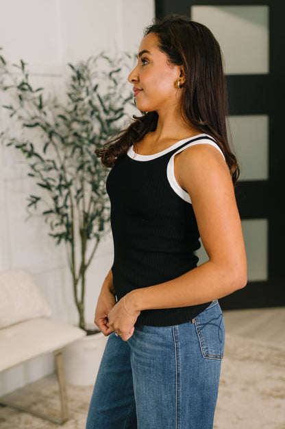 Woman wearing a black tank top with white trim and blue jeans standing indoors.