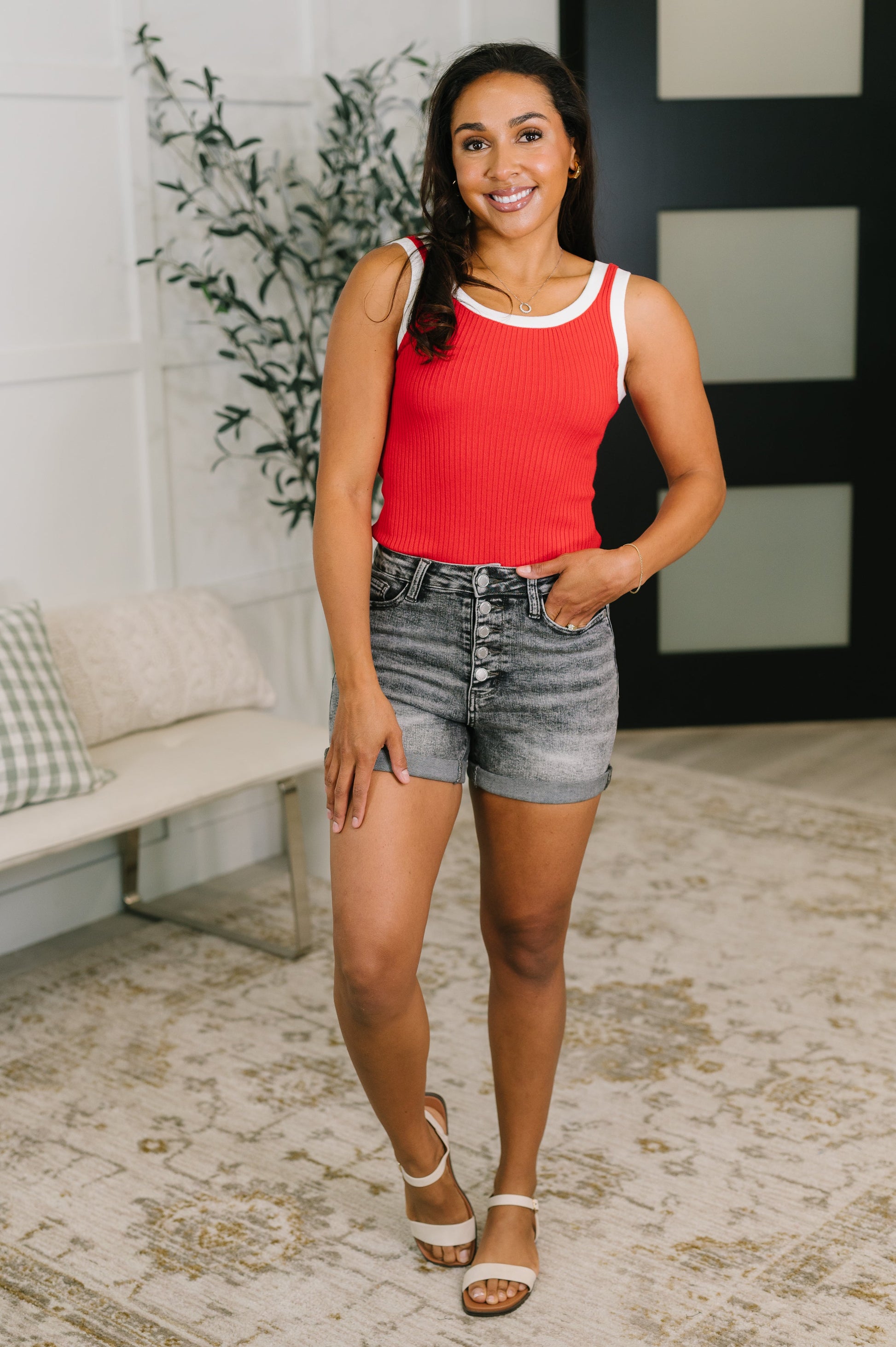 Woman wearing a red tank top and denim shorts standing in a room with a plant and bench.