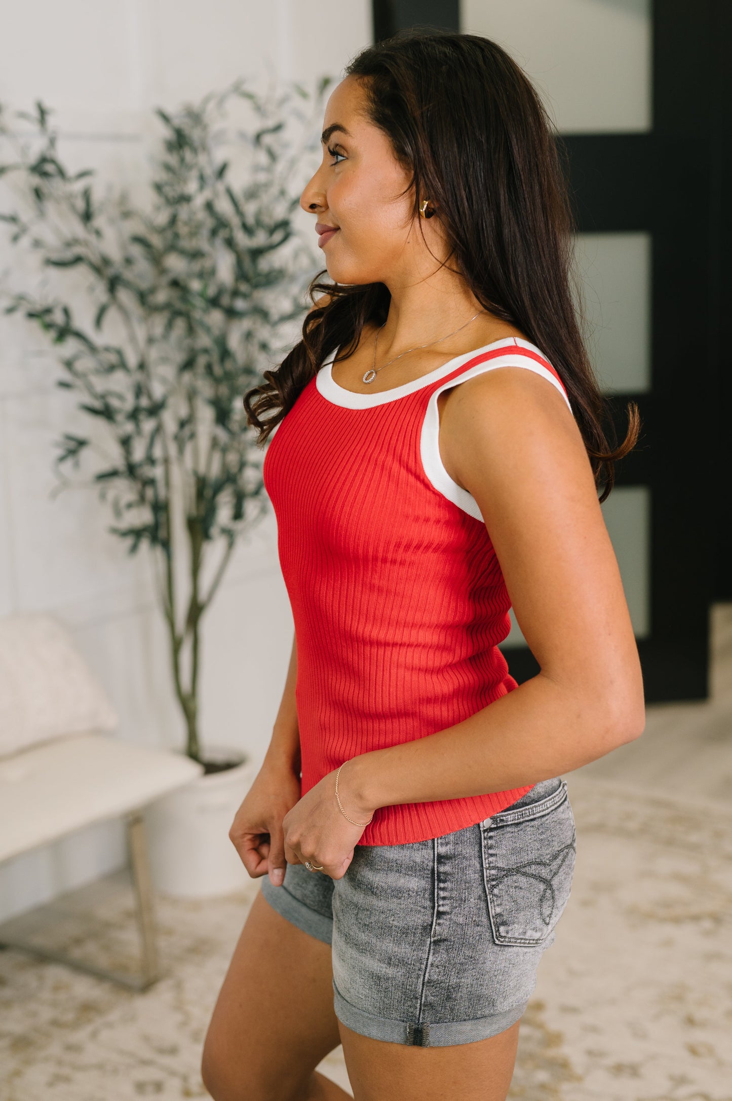 Woman wearing a red tank top and denim shorts standing indoors with a plant in the background