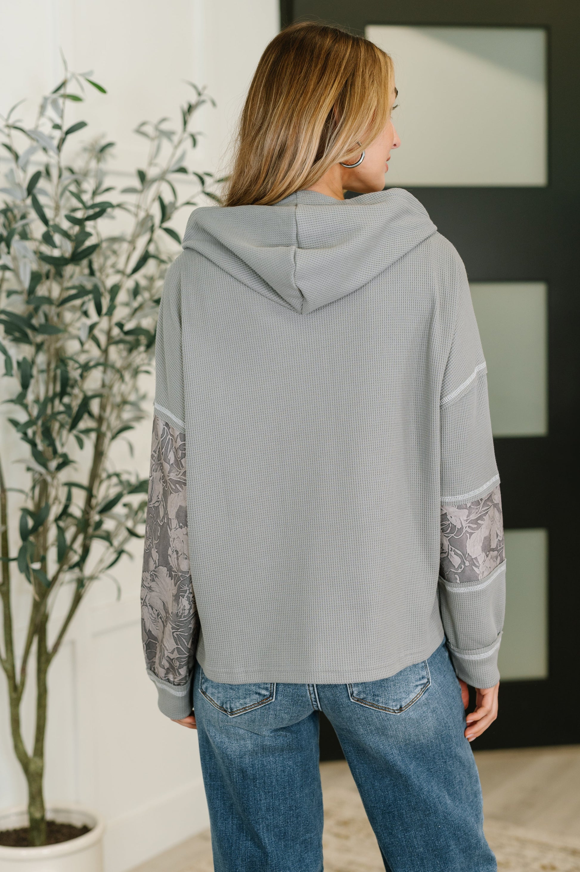 Person wearing a gray hoodie with lace sleeves in a room with a plant and framed picture.