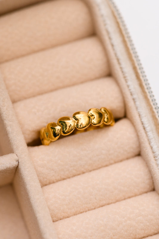 A gold-plated stainless steel ring with a hearts on hearts design, displayed in a soft pink jewelry box.