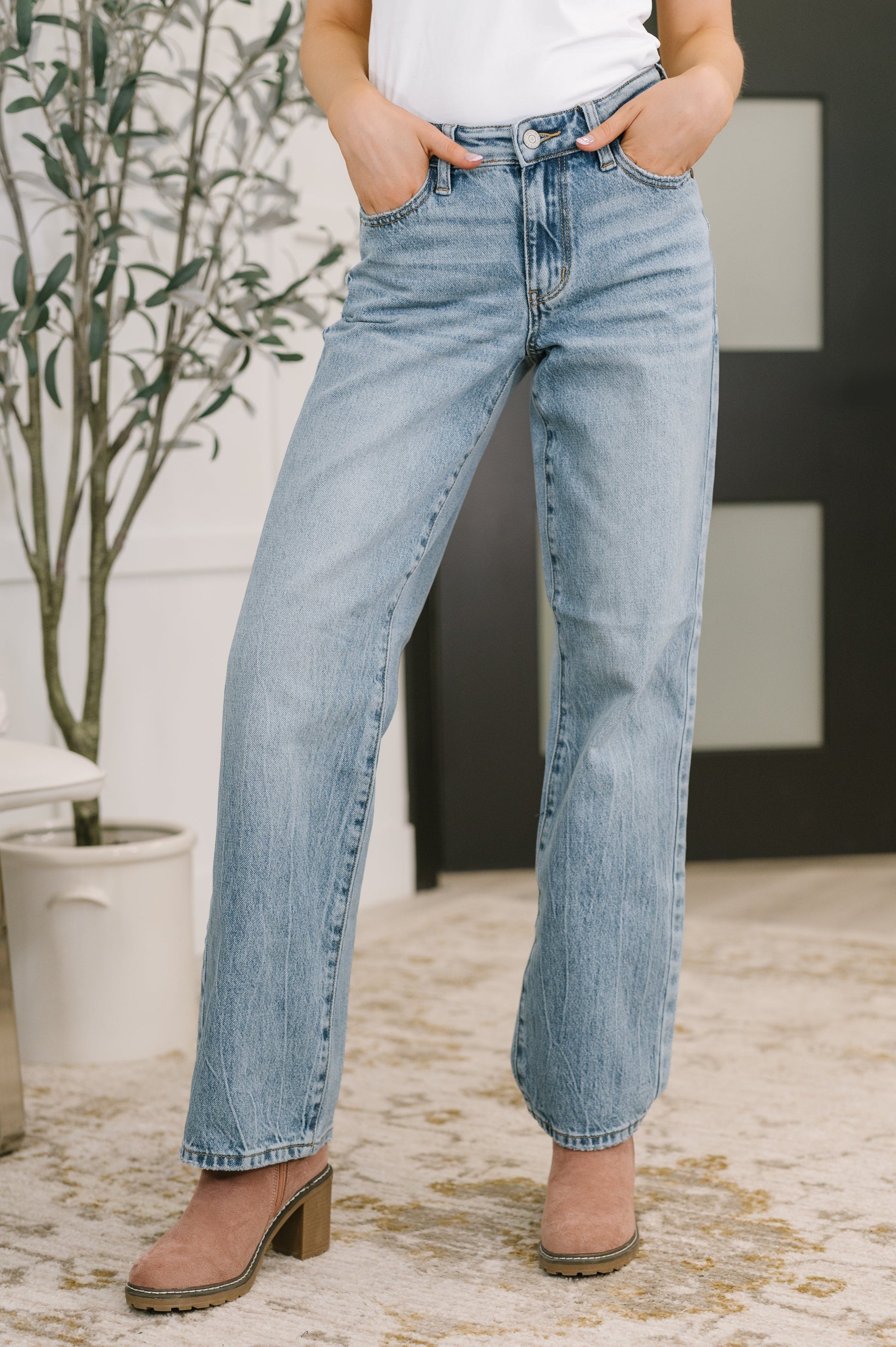 Person wearing light blue jeans standing indoors with a plant in the background