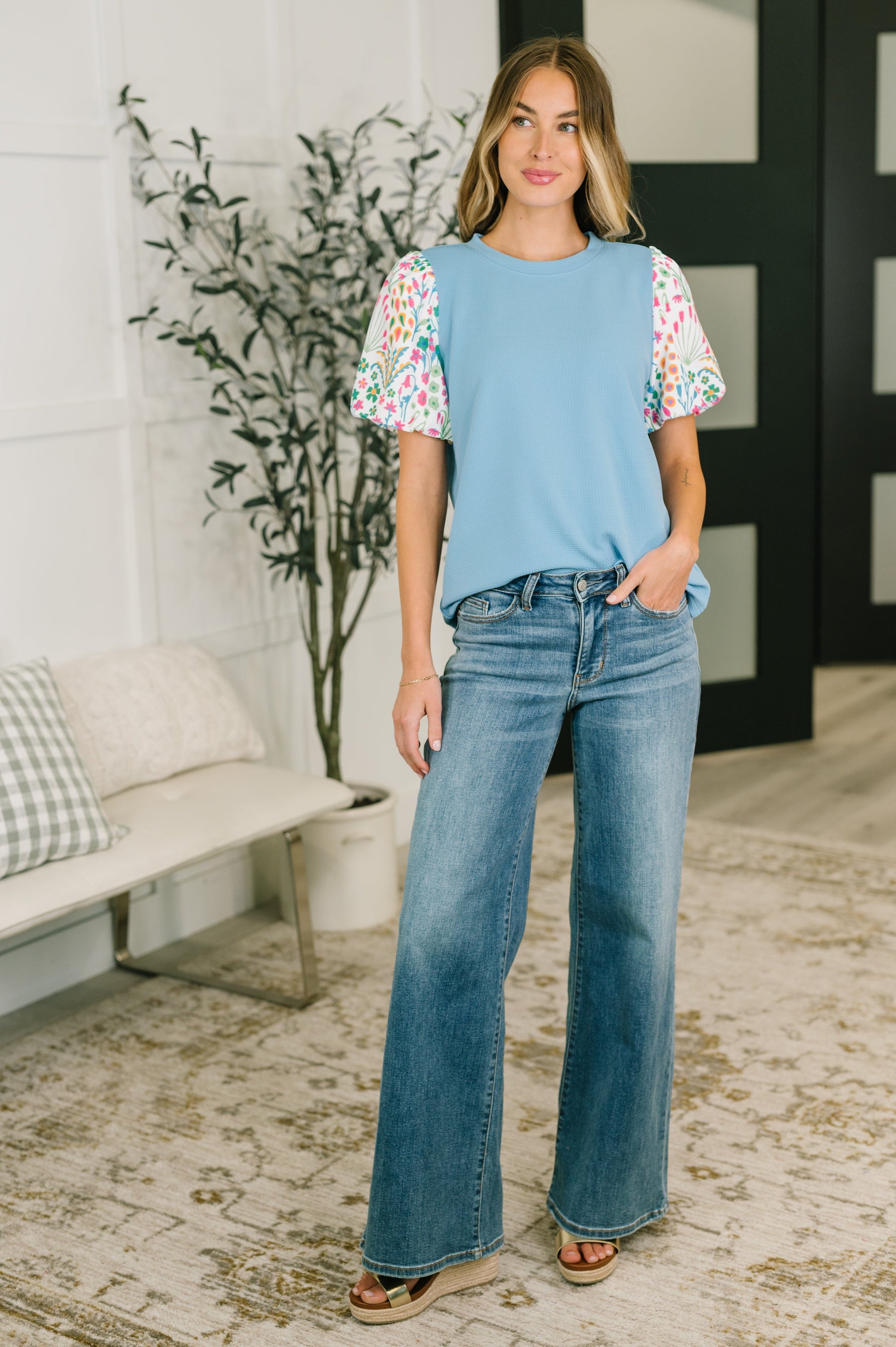 Woman wearing a light blue top with floral sleeves and jeans standing in a room with a plant and bench.
