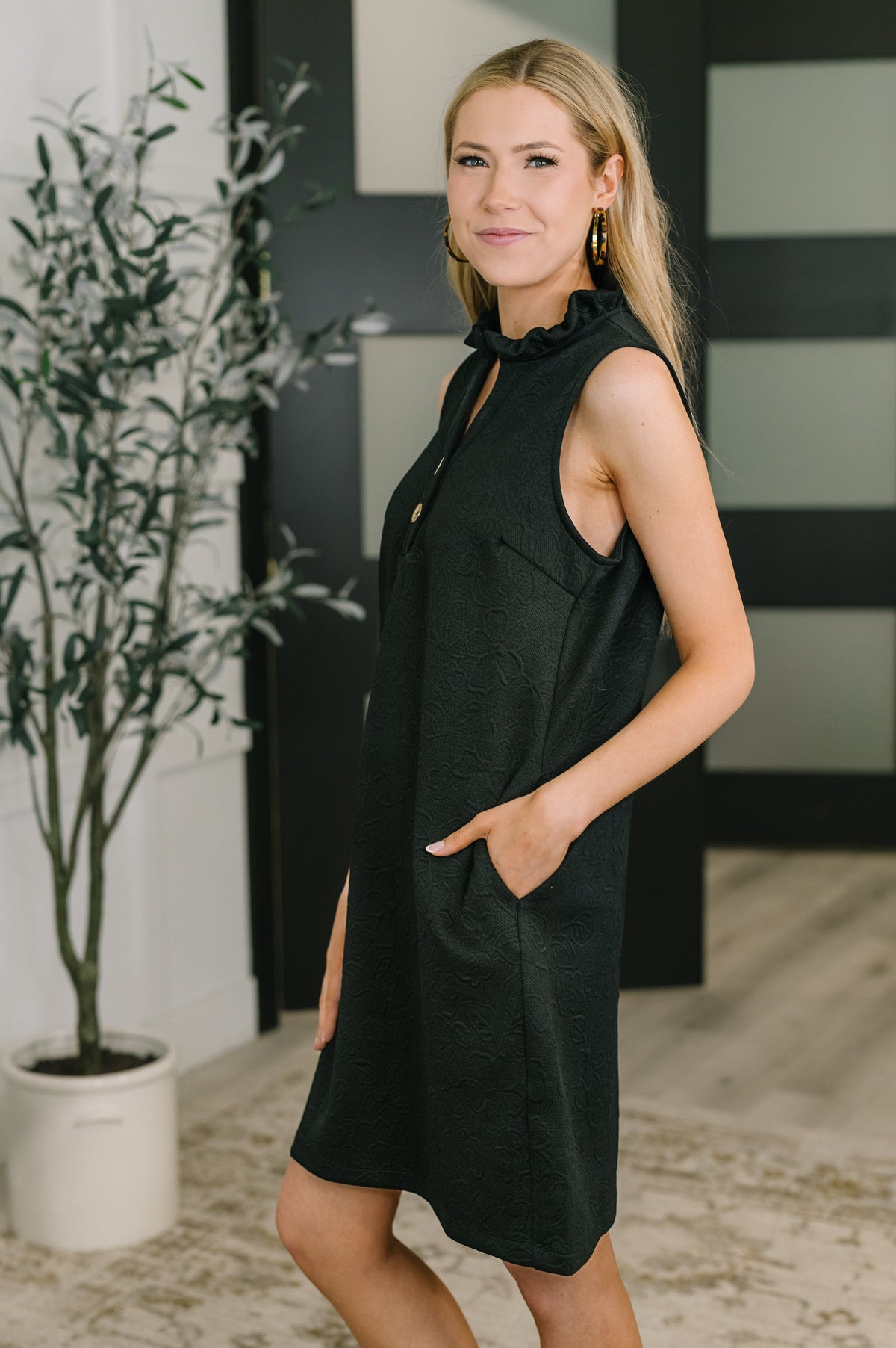 Woman wearing a black sleeveless dress with a plant and striped wall in the background