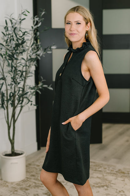 Woman wearing a black sleeveless dress with a plant and striped wall in the background