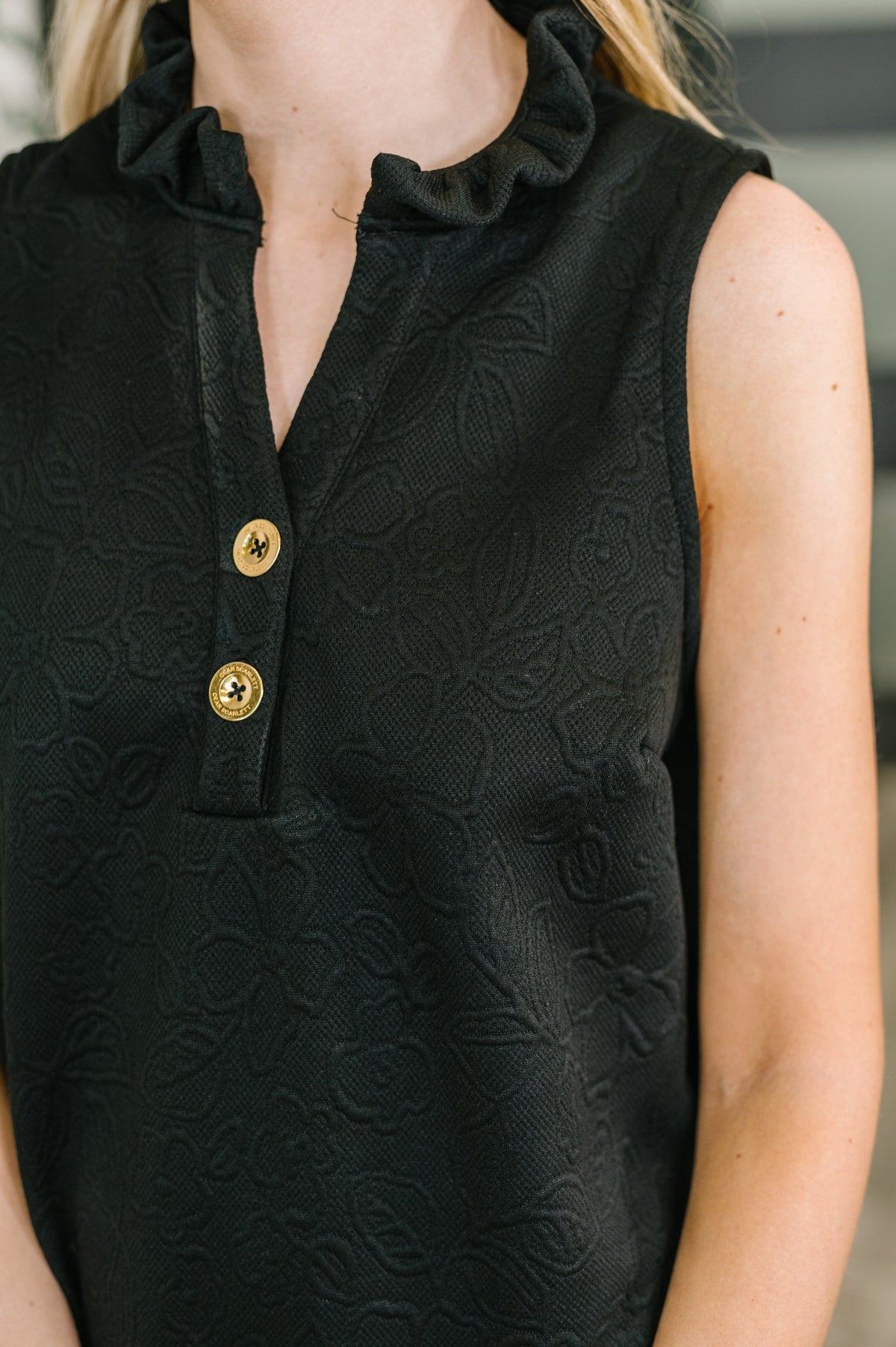 Black sleeveless top with gold buttons worn by a person, blurred background