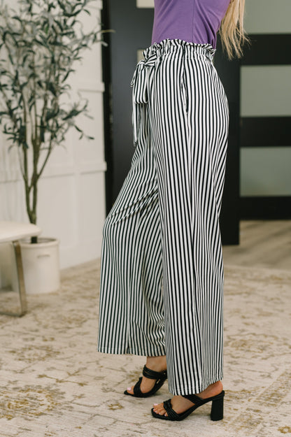 A person wearing high-waisted, wide-leg striped pants with black and white vertical stripes, paired with a purple top and black heeled sandals, standing on a patterned rug in an indoor setting.