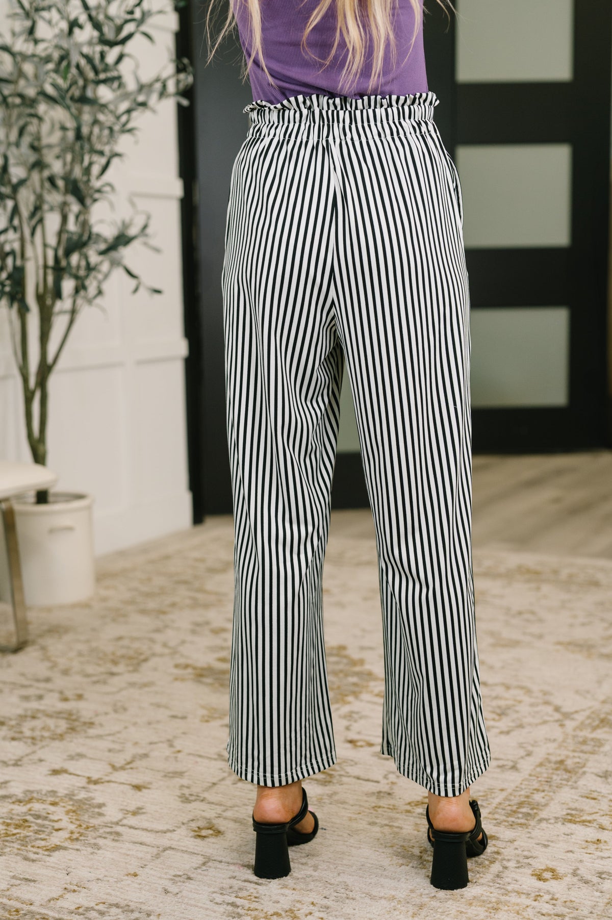 A person shown from the back wearing black and white vertically striped wide-leg cropped pants, a purple top, and black heeled sandals in an indoor setting with a patterned rug and potted plant.