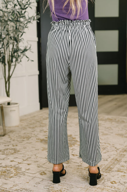A person shown from the back wearing black and white vertically striped wide-leg cropped pants, a purple top, and black heeled sandals in an indoor setting with a patterned rug and potted plant.