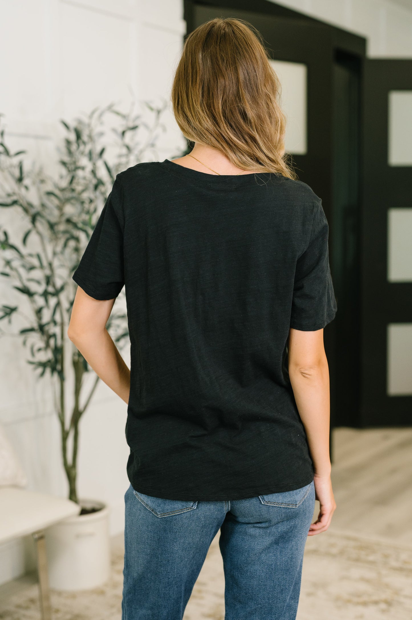 Person wearing a black t-shirt and blue jeans standing in a room with a plant and door in the background.