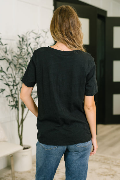 Person wearing a black t-shirt and blue jeans standing in a room with a plant and door in the background.