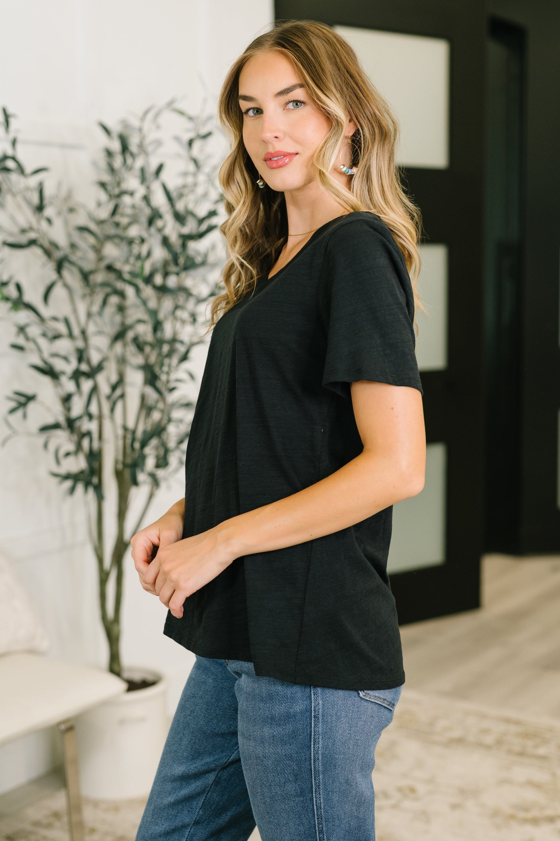 Woman wearing a black t-shirt and blue jeans standing indoors with a plant in the background