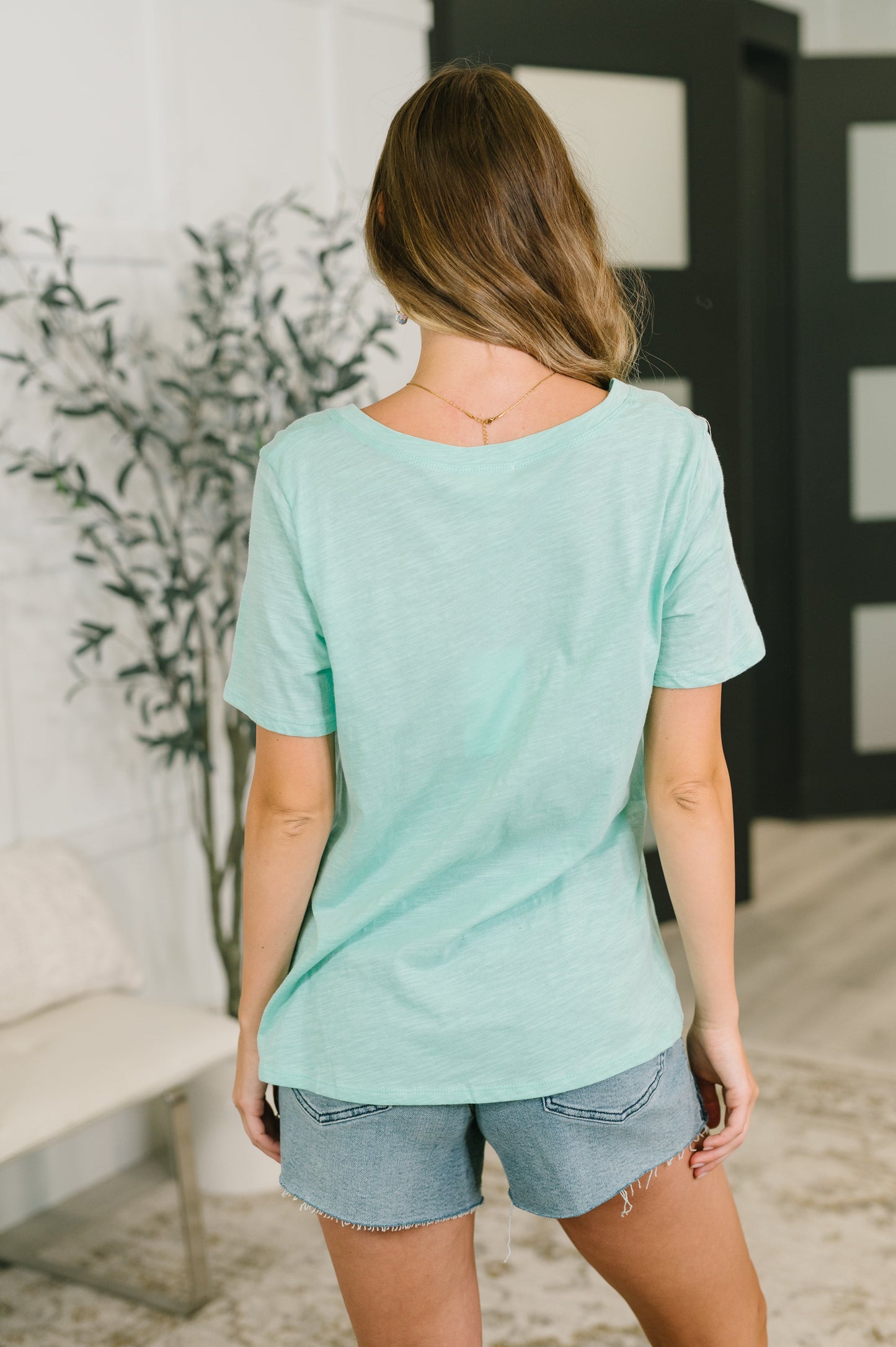 A woman viewed from behind wearing a mint green short-sleeve t-shirt and denim shorts in a modern indoor setting.