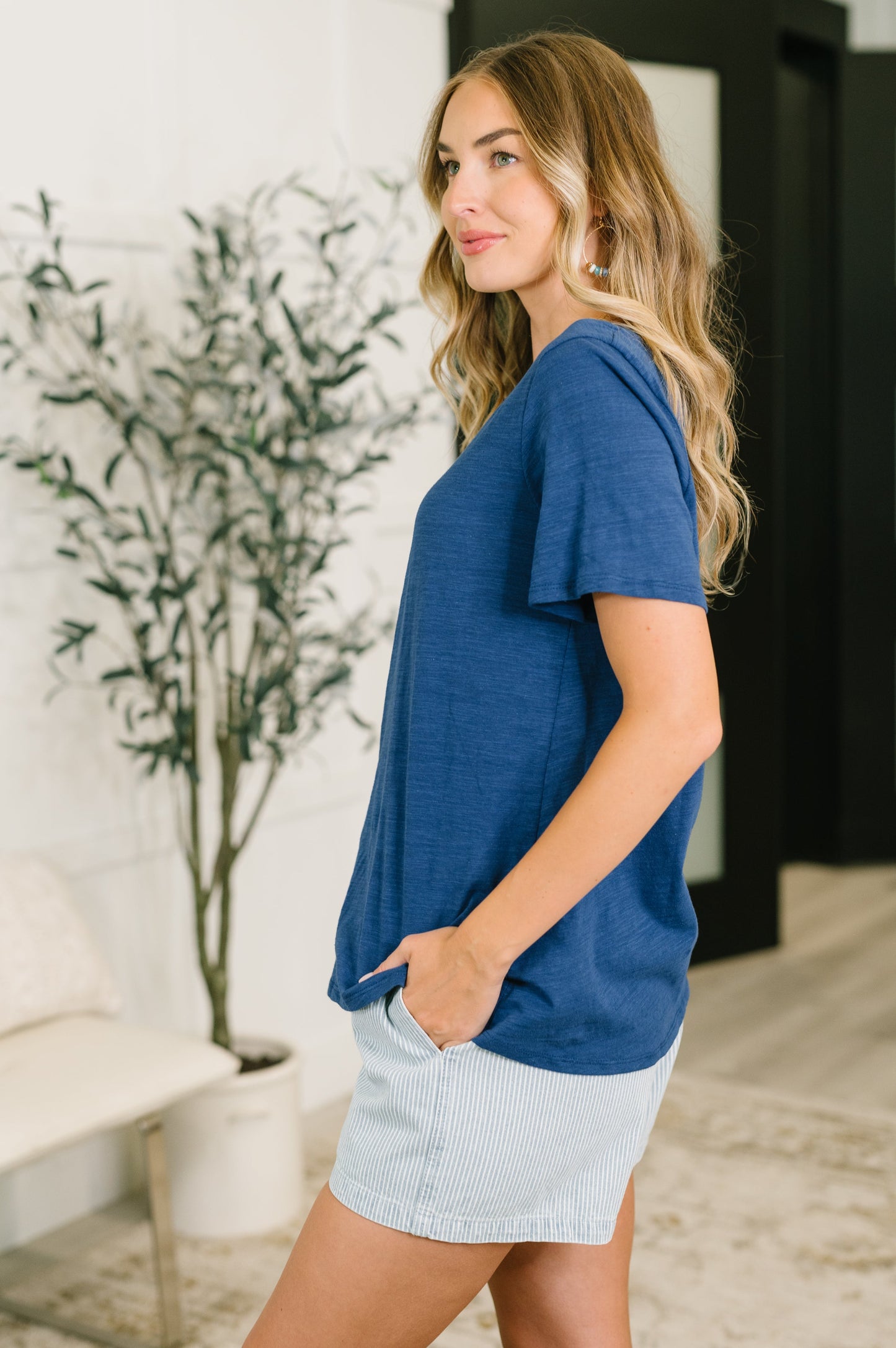 Woman wearing a blue top and light shorts indoors with a plant in the background