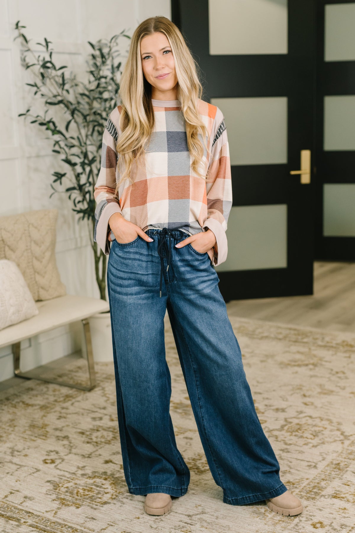A woman stands indoors wearing a long-sleeve plaid top in peach, gray, and white with wide-leg blue jeans, standing on a patterned rug in front of a black door.