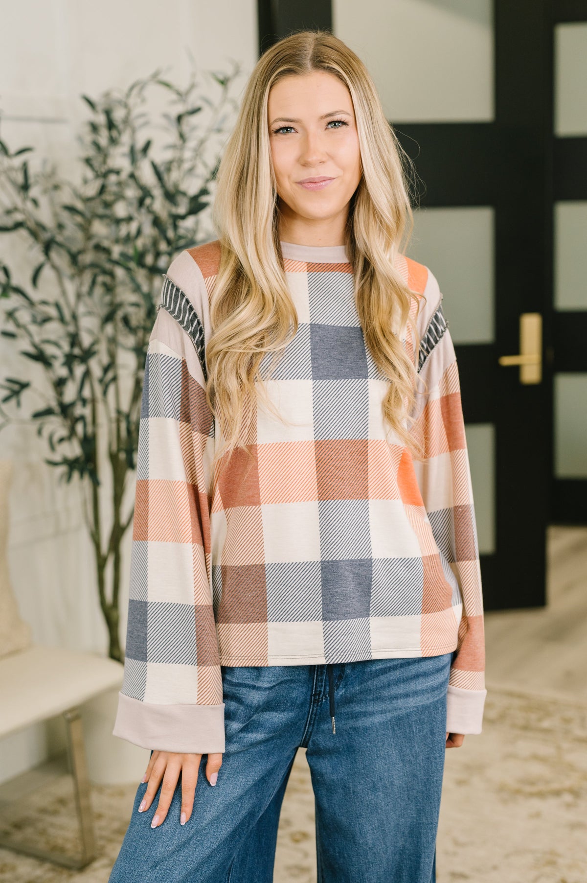 A woman modeling a long-sleeve plaid sweater with a check pattern in peach, gray, and cream, paired with blue jeans in an indoor setting.
