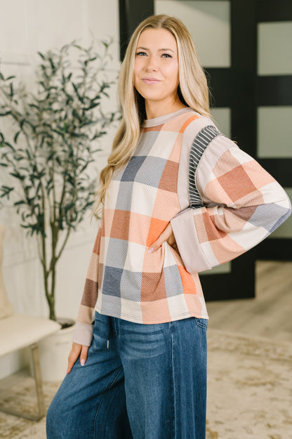A woman modeling a long-sleeve plaid sweater in peach, gray, and cream, paired with blue jeans in a modern indoor setting.