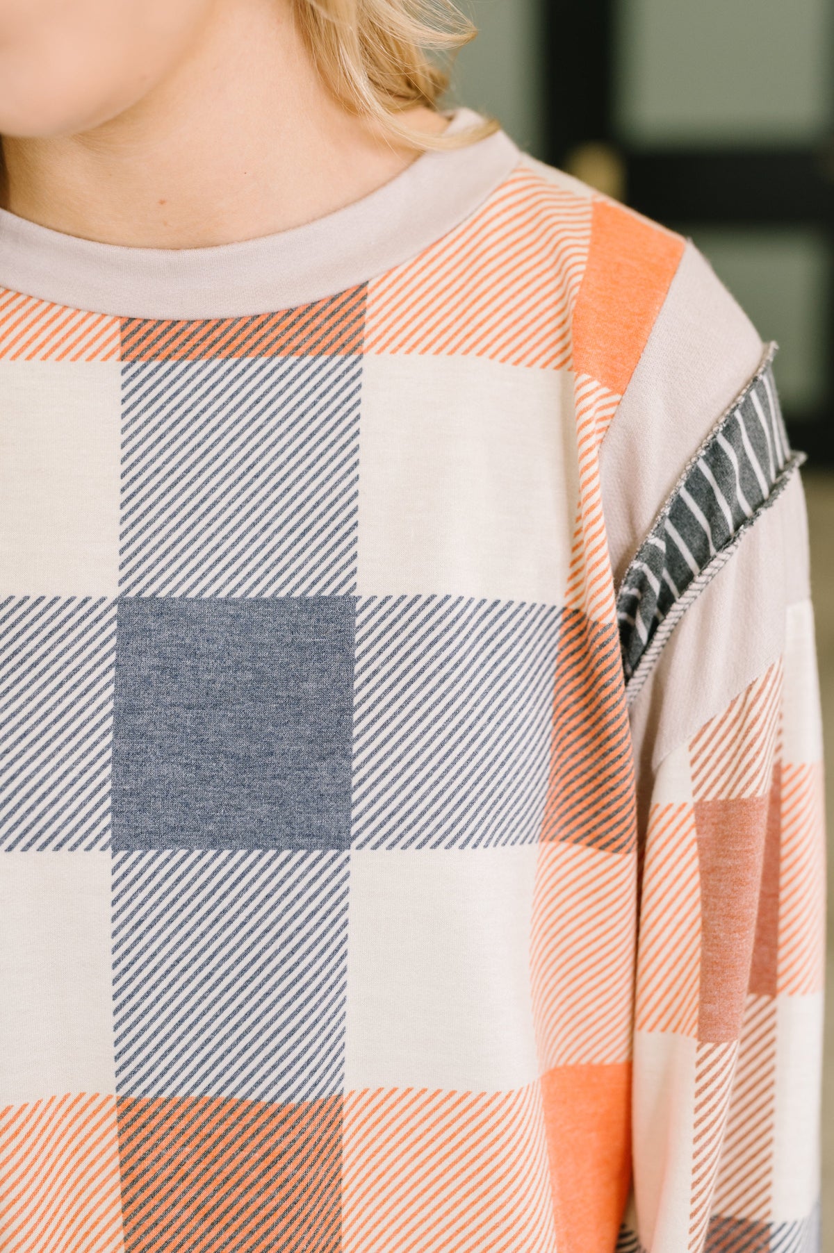 Close-up of a person wearing a long-sleeve plaid top with peach, gray, and off-white check blocks, plus a striped and textured sleeve detail.