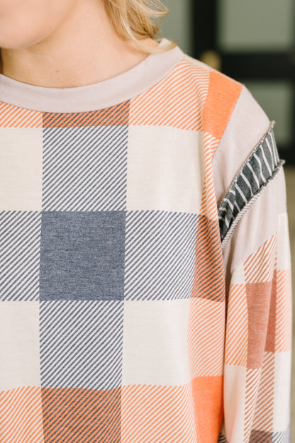 Close-up of a person wearing a long-sleeve plaid top with peach, gray, and off-white check blocks, plus a striped and textured sleeve detail.