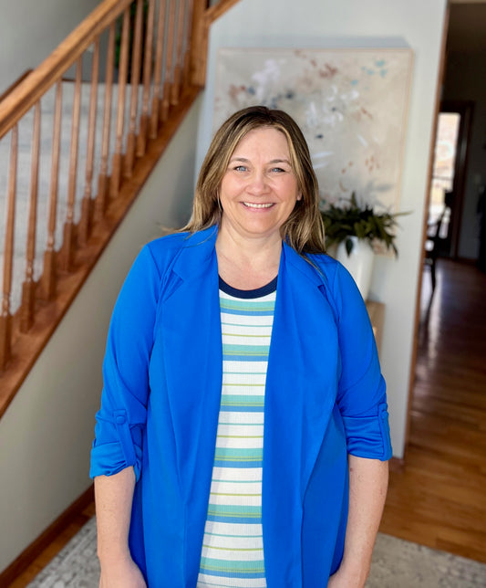Woman in a blue jacket standing in a home interior with a staircase and artwork in the background.