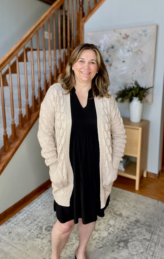 Woman wearing a beige cardigan over a black dress in a home setting with a staircase and artwork in the background.