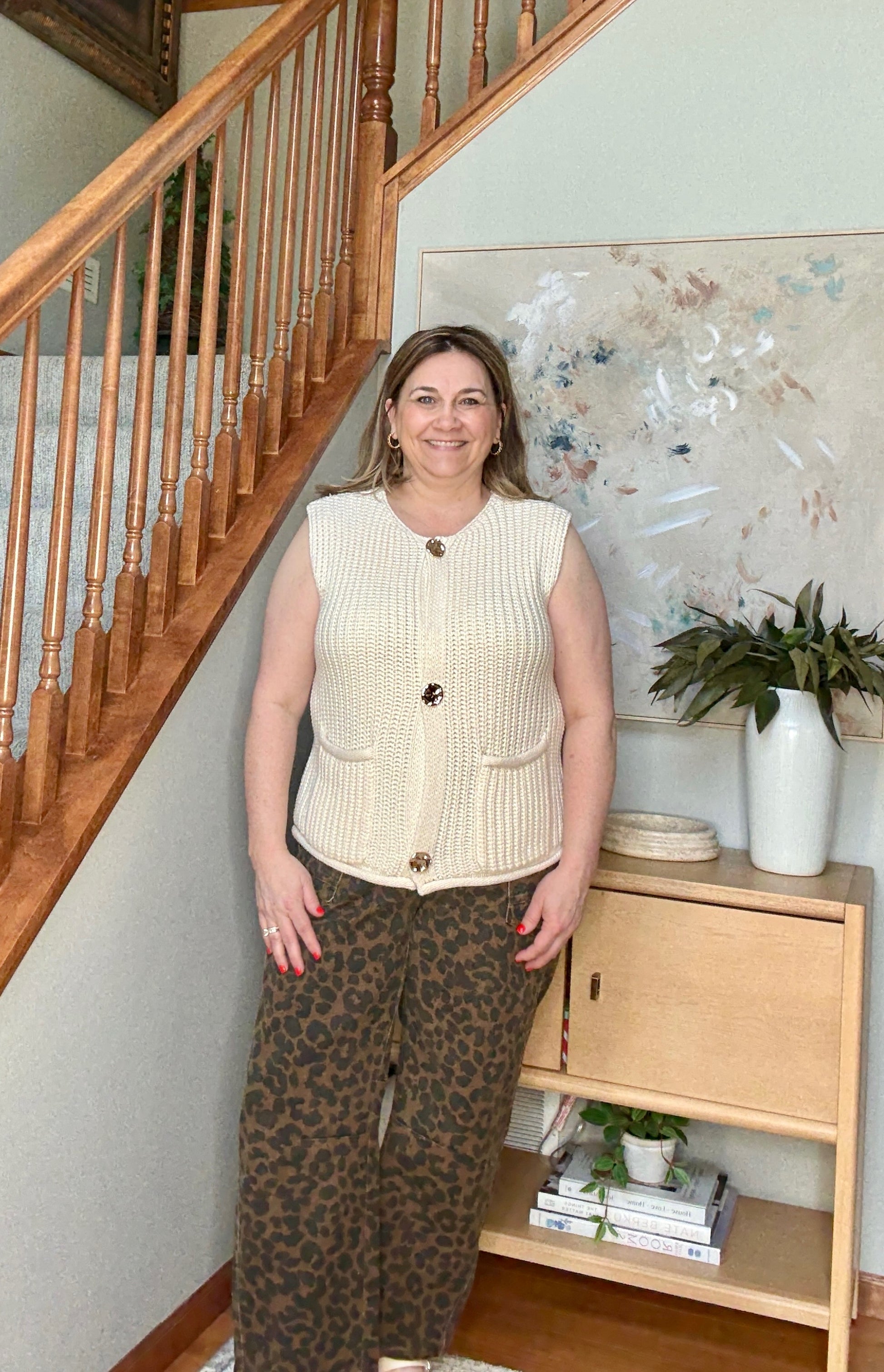 Woman standing in a home setting with wooden staircase and bookshelf.