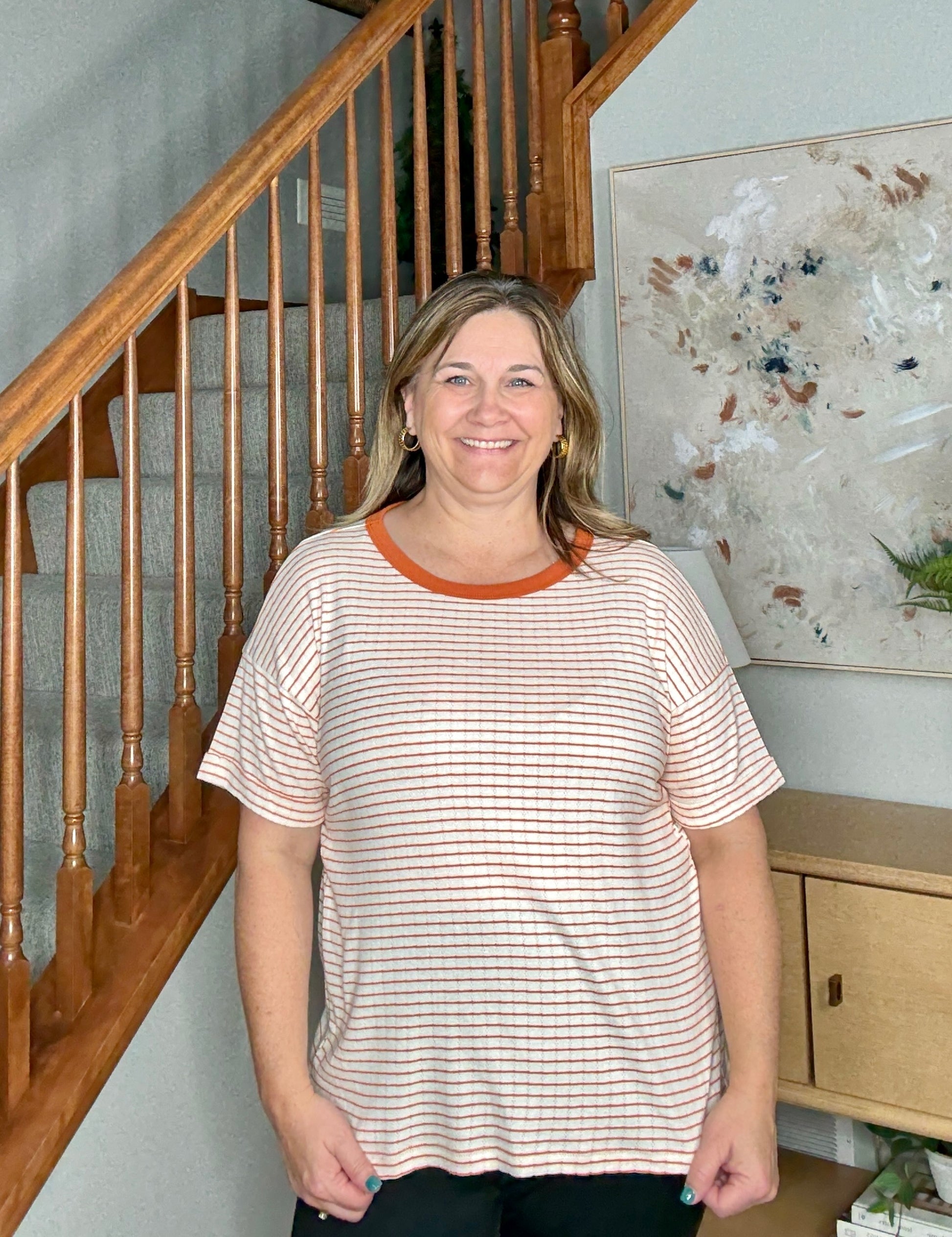 Woman standing in a home setting with wooden staircase and artwork on the wall.