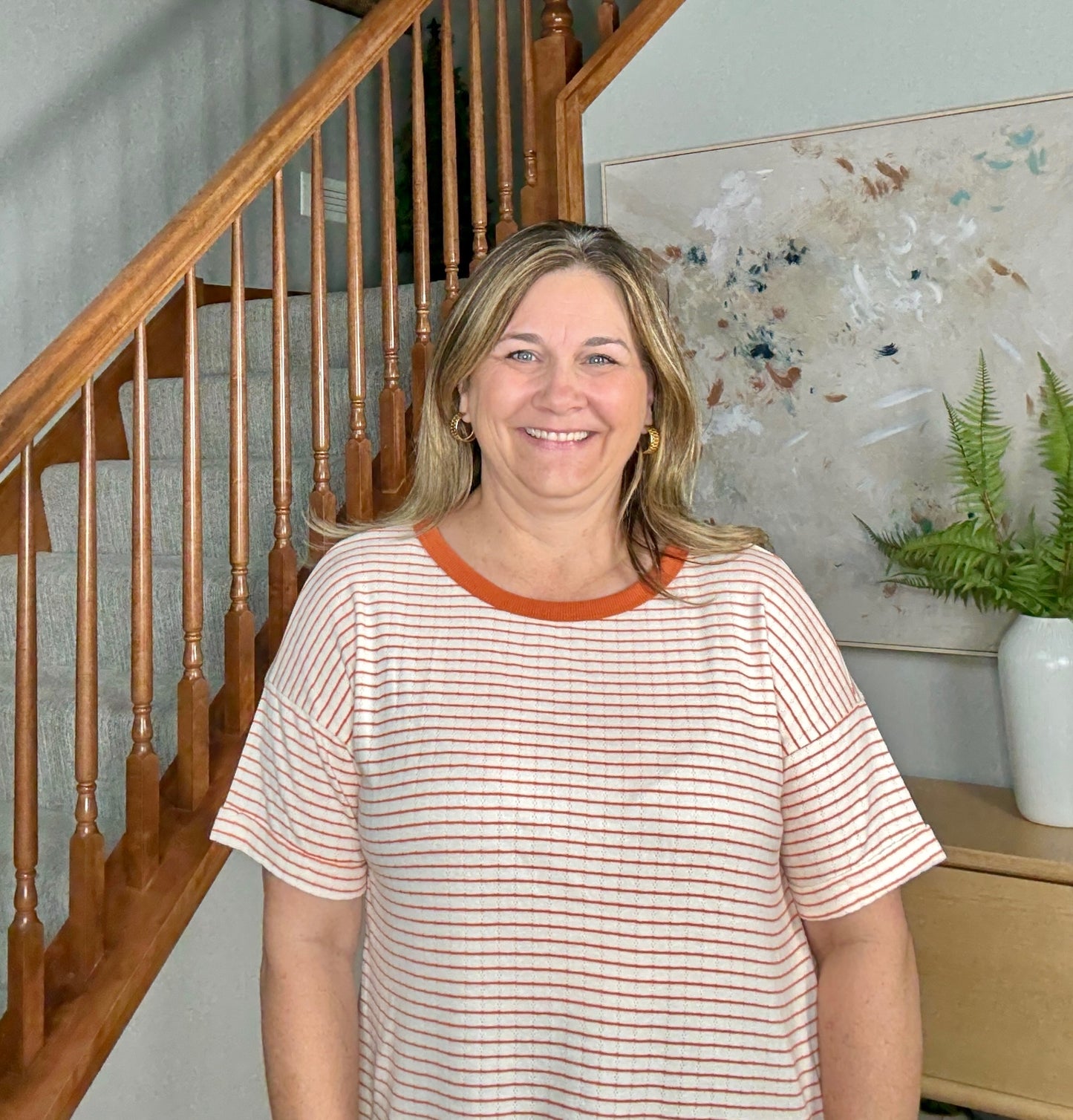 Woman standing in a home setting with wooden staircase and decorative elements.