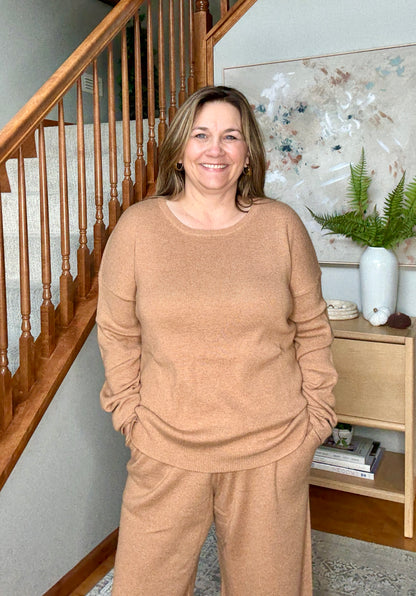 Woman in a beige sweater and pants standing in a home interior with wooden staircase and decor.