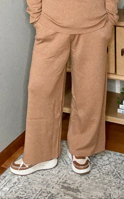 Woman in a beige outfit standing in a home setting with a staircase and decorative items.