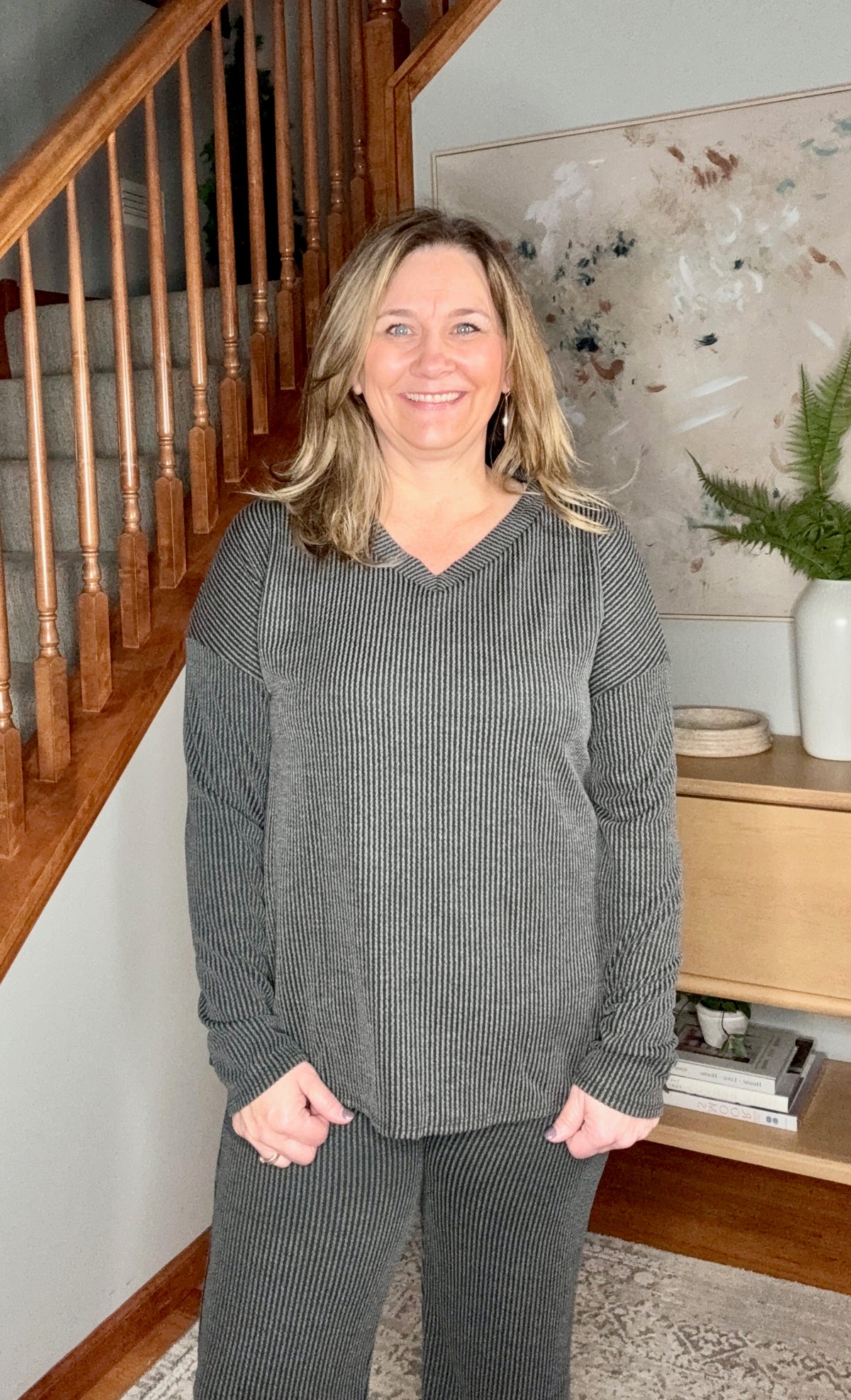 Woman in a gray sweater standing in a home setting with a staircase and decor.