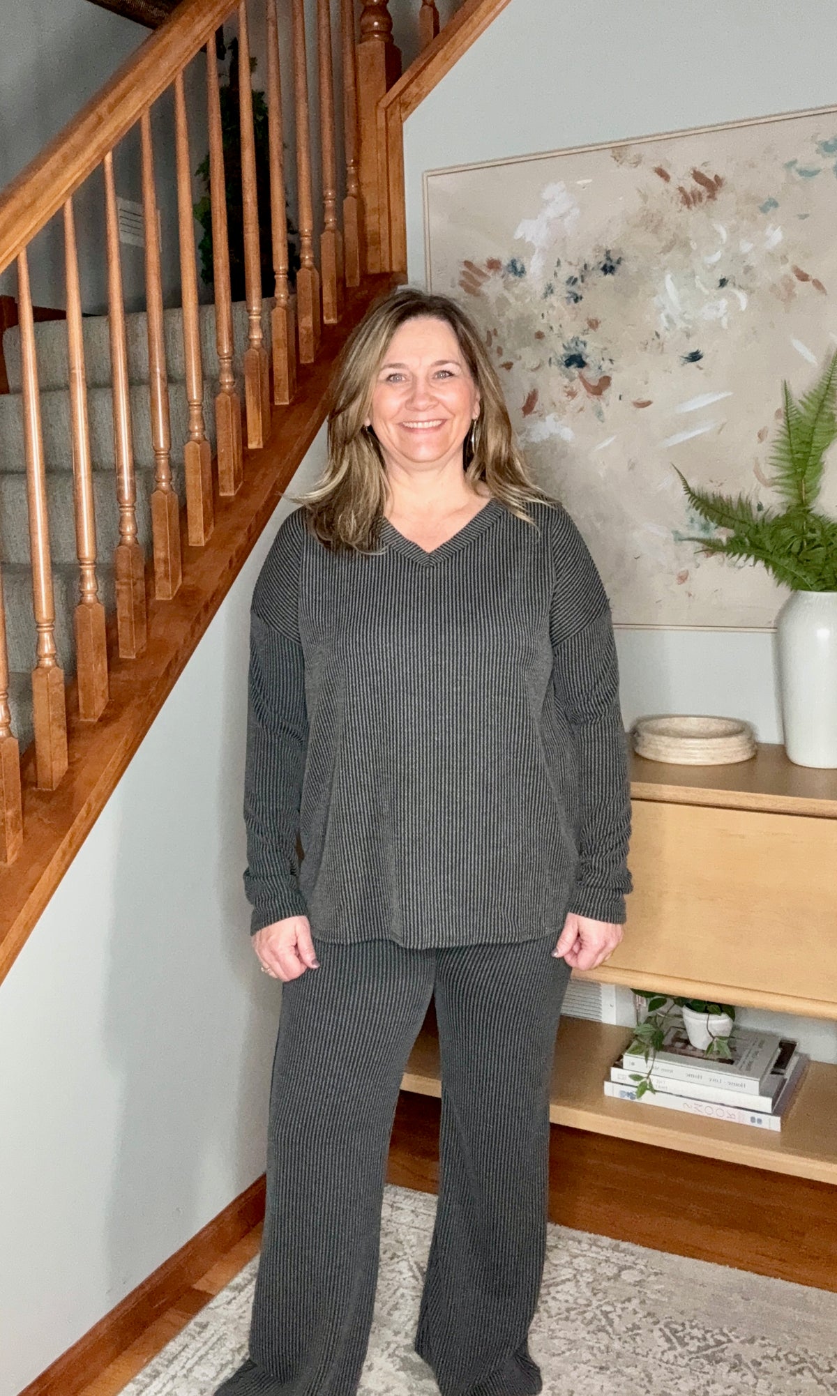 Woman in gray outfit standing in a home interior with wooden staircase and decor.