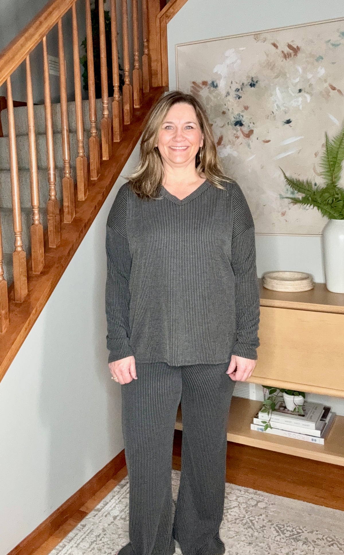 Woman in a gray outfit standing in a home interior with wooden staircase and decor.