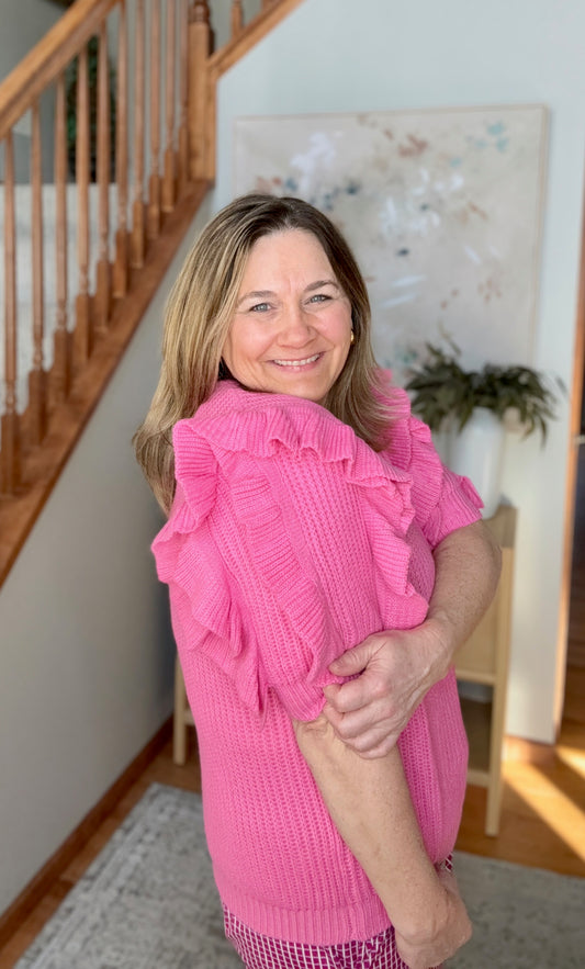 Woman wearing a pink ruffled blouse in a home setting with wooden staircase and decor.