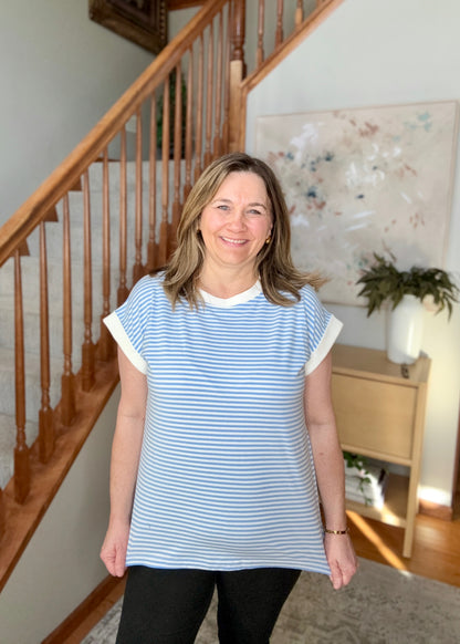 Woman wearing a blue and white striped shirt standing in a home setting with a staircase and artwork in the background.