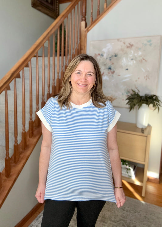 Woman wearing a blue and white striped shirt standing in a home setting with a staircase and artwork in the background.