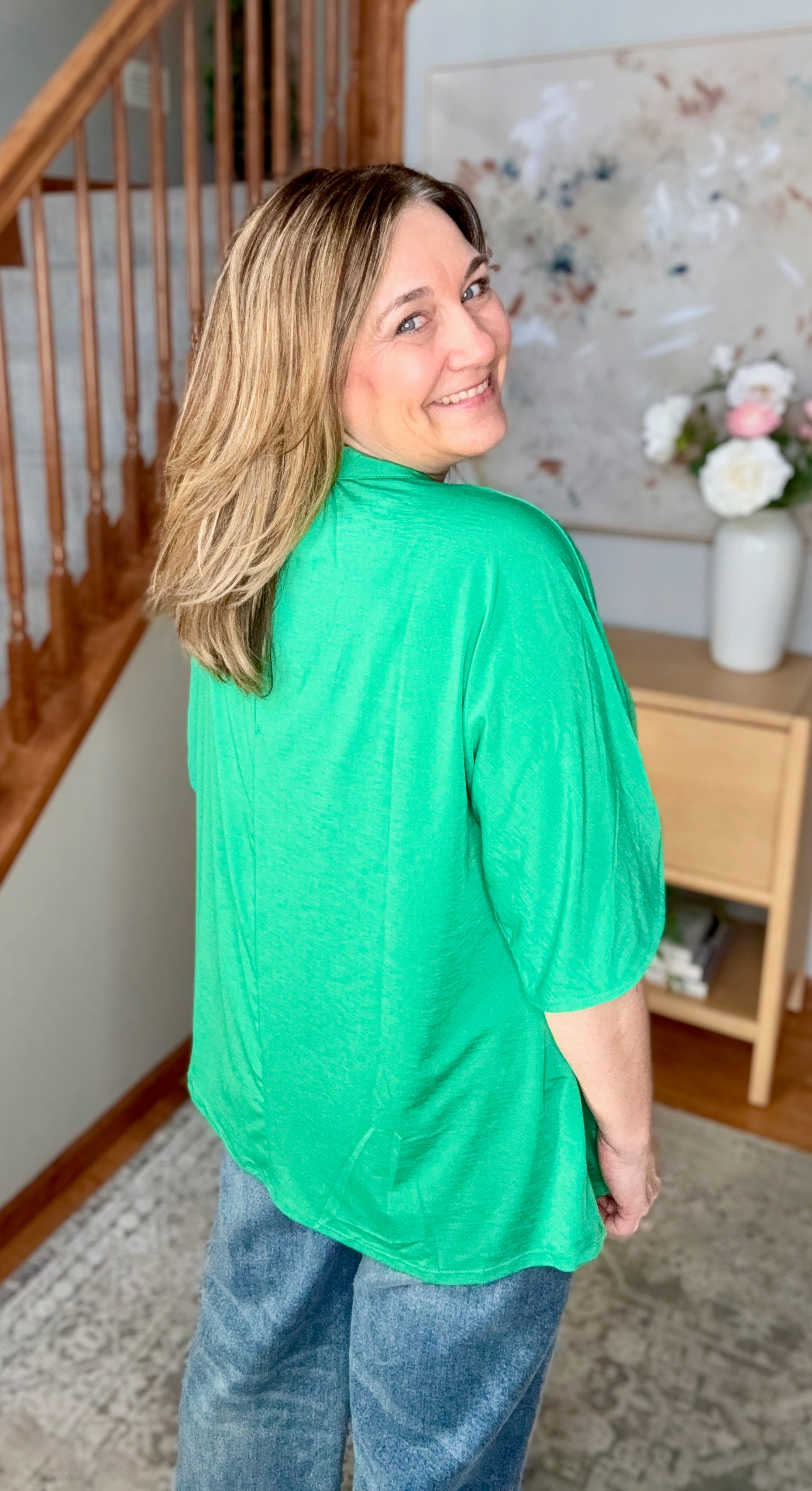 Woman wearing a green top and blue jeans standing in a home setting with a staircase and floral arrangement in the background.