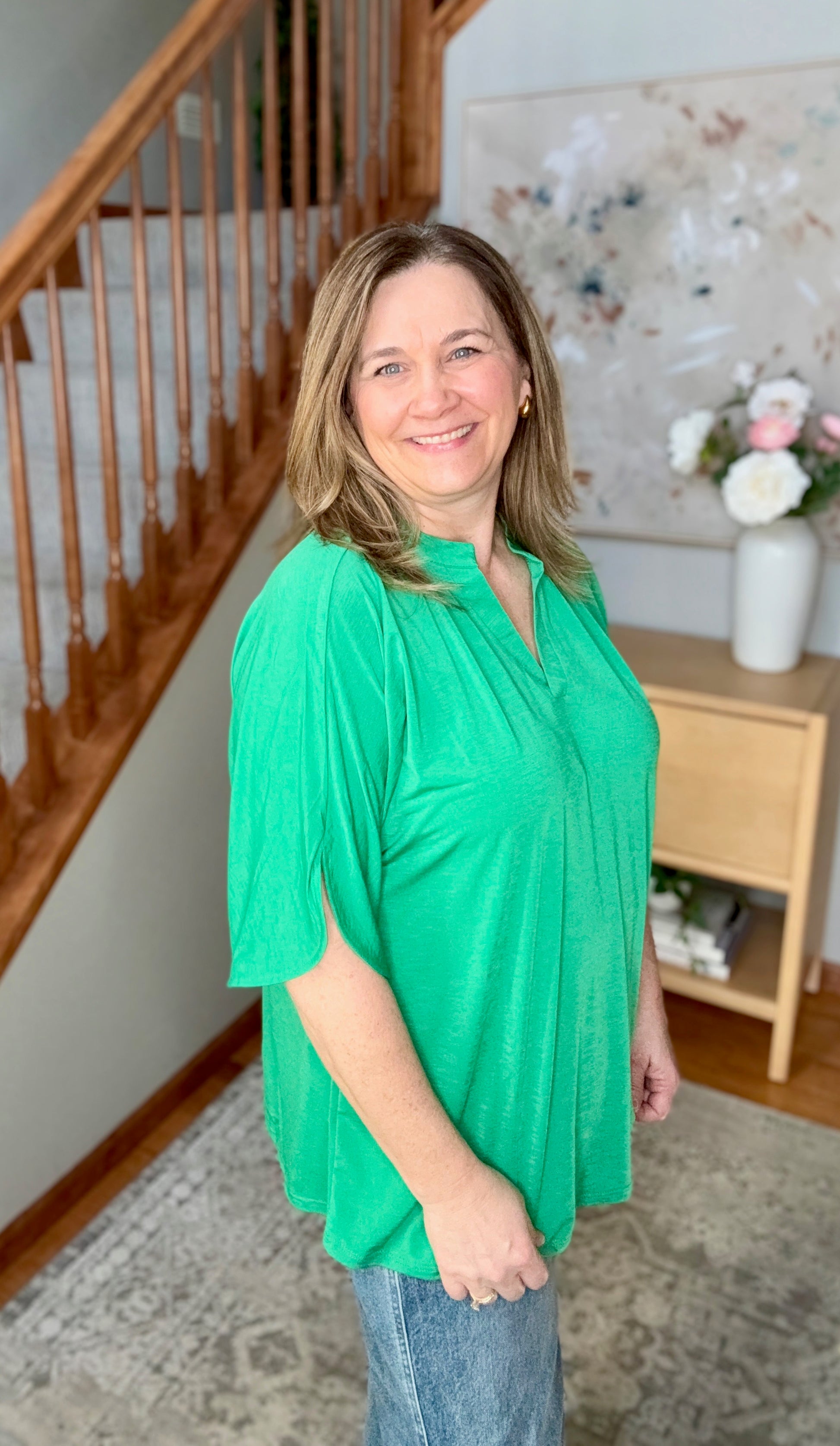 Woman in a green top standing in a home setting with a staircase and decor.
