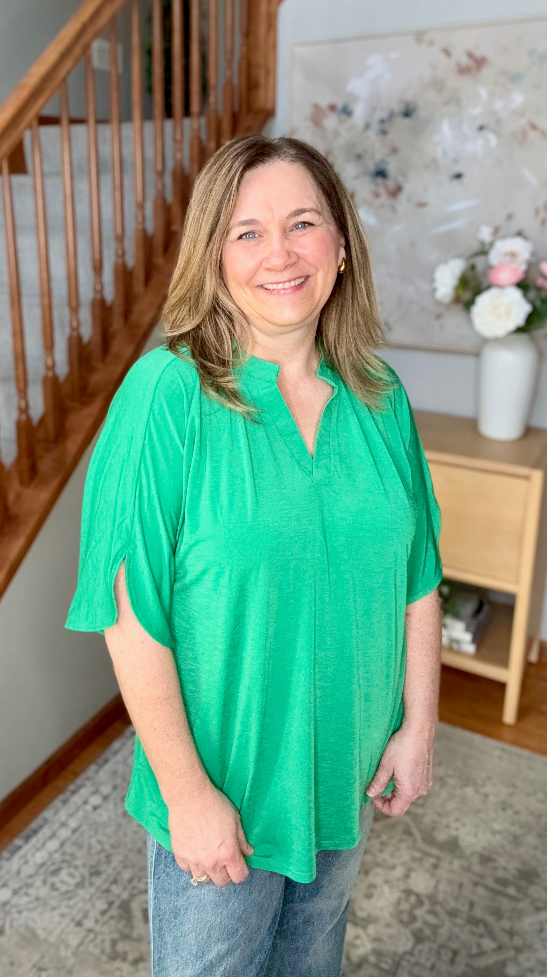 Woman wearing a green blouse standing in a home setting with a staircase and decorative items.