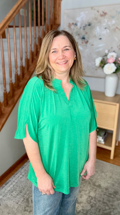 Woman wearing a green blouse standing in a home setting with a staircase and decorative items.